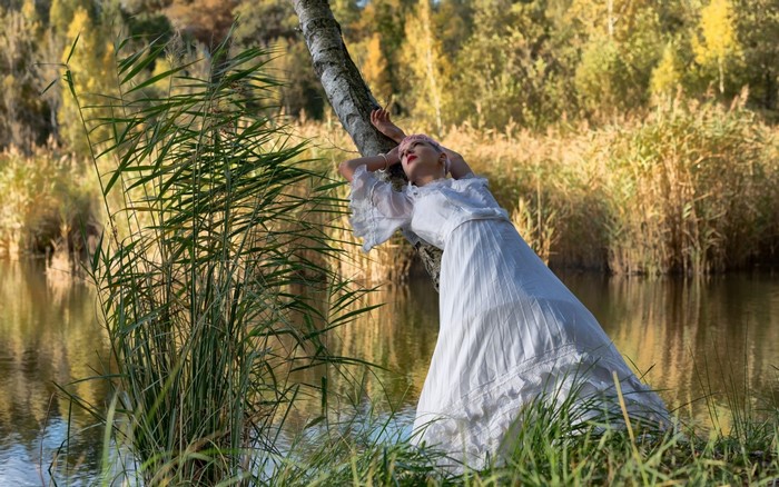 Woman in white dress leaning on a tree by a lake