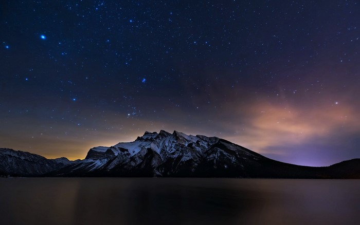 Snowy mountains under a starry night sky reflected in a lake