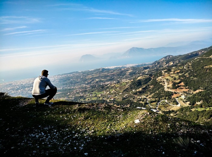 Man crouching on a hill overlooking a coastal city and mountains