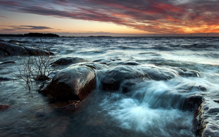 Waves crashing on rocks at sunset with wind turbines on the horizon