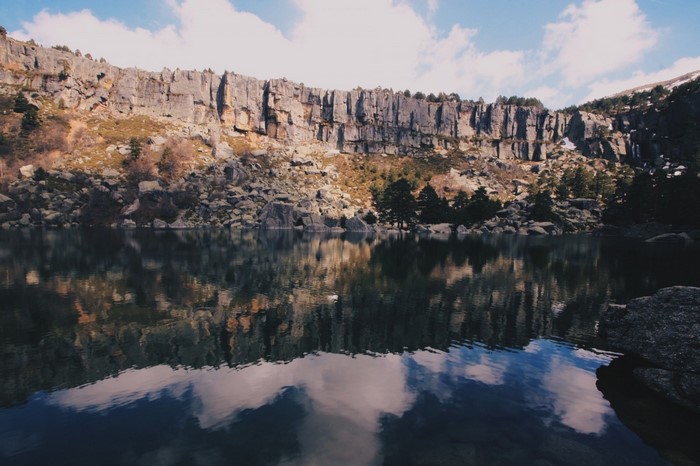 Lake reflecting a rocky cliff face and trees under a cloudy sky
