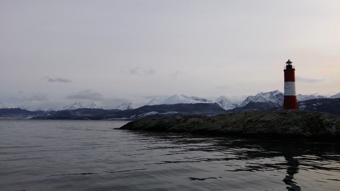 Red and white lighthouse on rocky shore with snow-capped mountains