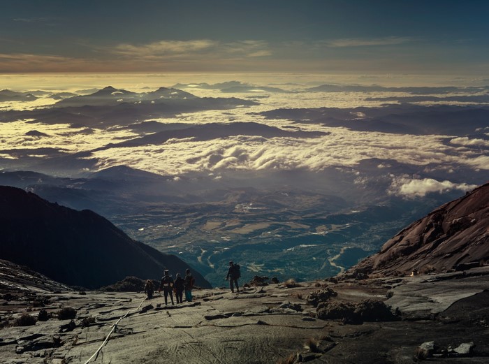 Hikers on a rocky mountain slope above a cloud-filled valley at sunrise