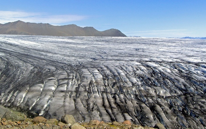 Glacier with dark streaks and mountains under blue sky