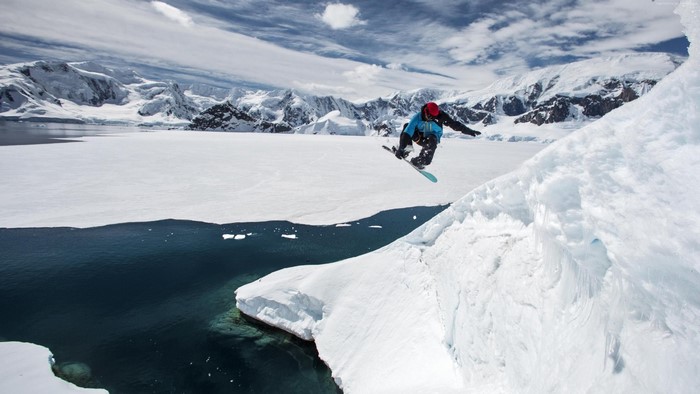 Snowboarder jumping over icy water with snowy mountains