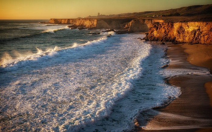 Waves crash on a sandy beach with cliffs at sunset