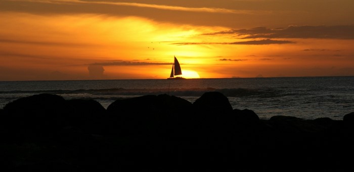 Sailboat on the ocean at sunset with dark rocks in foreground