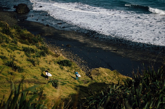 Two surfers walking down a grassy hill towards a rocky beach and ocean
