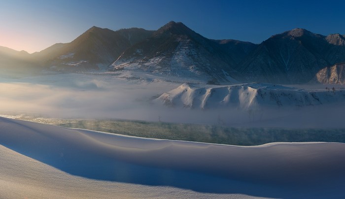 Snowy mountain valley with mist over a river at sunrise