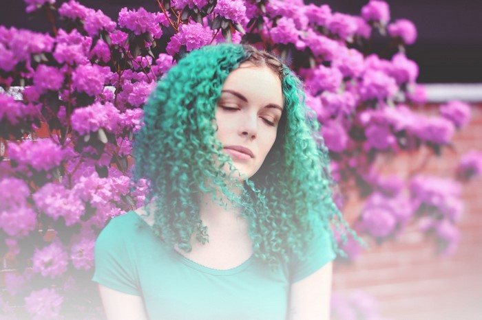 Woman with teal curly hair in front of purple flowers
