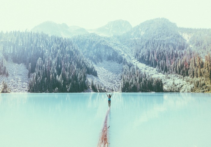 Woman on log in turquoise lake with mountains