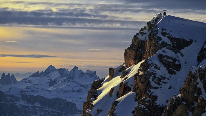 Snowy mountain peaks at sunset with a cross on the summit