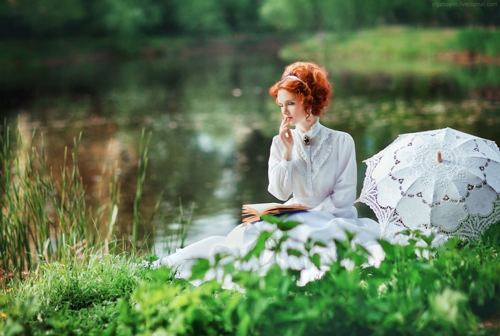 Redhead woman in white dress reading by a pond with umbrella