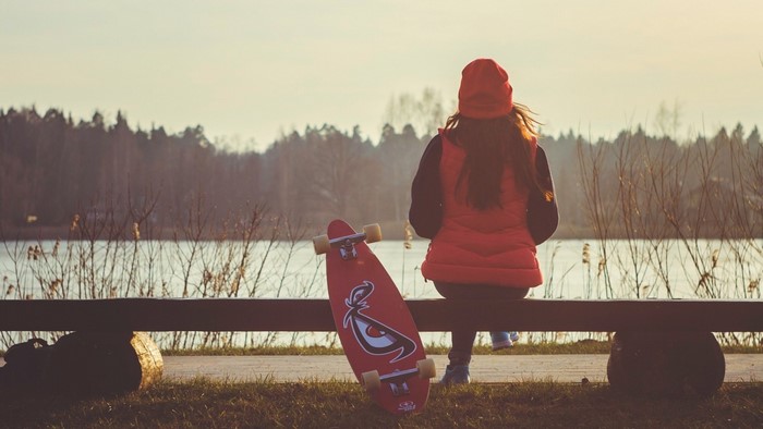 Girl in red sitting on bench with skateboard by lake
