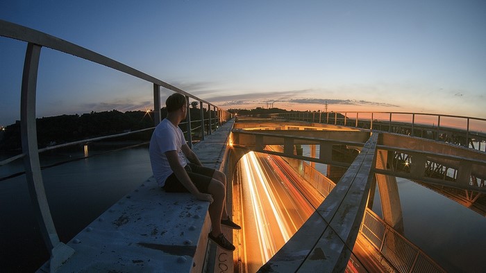 Man sitting on a bridge overlooking car light trails at sunset