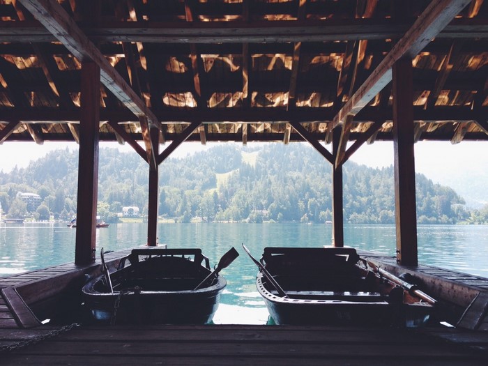 Two boats docked under a wooden boathouse with a lake and forest background