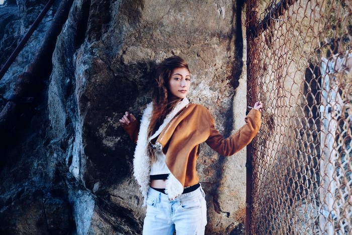 Woman in brown jacket and jeans by a rock and fence