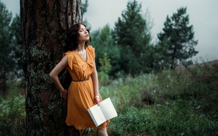 Woman in orange dress leaning against tree holding a book