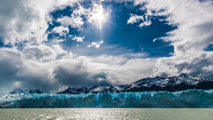Glacier and snow-capped mountains under a cloudy sky with sun