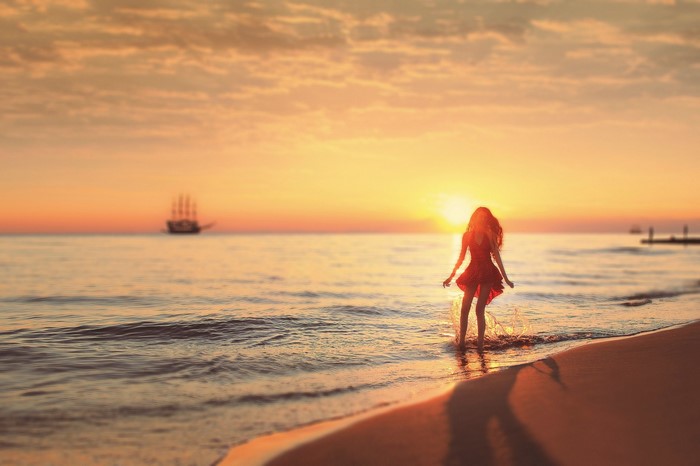 Woman in red dress on beach at sunset with sailboat