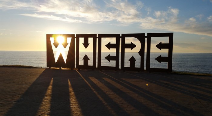 Large metal letters spelling WHERE with arrows at sunset by the sea