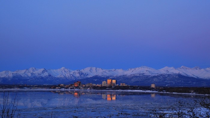 Cityscape reflected in water with snow-capped mountains at dawn