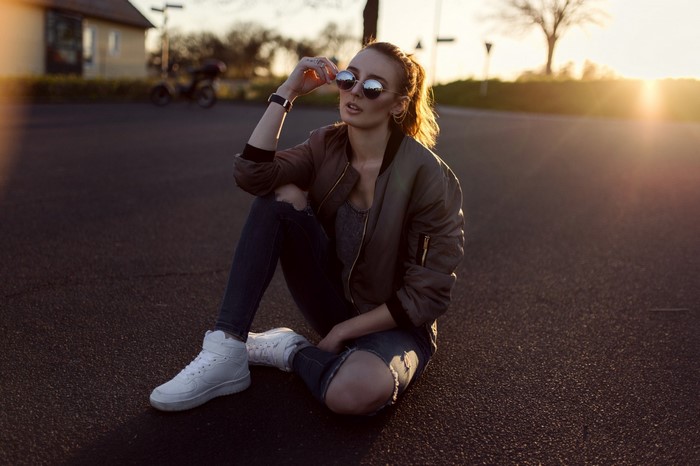 Woman in sunglasses and bomber jacket sitting on road at sunset