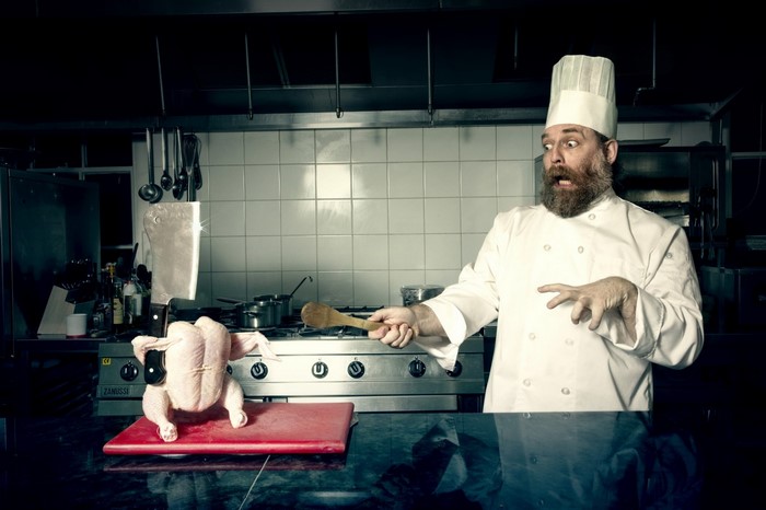 Chef with a surprised expression holding a wooden spoon near a raw chicken