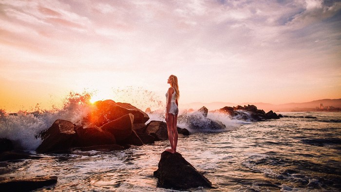 Woman standing on rock at sunset with waves crashing