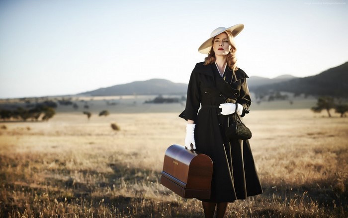 Kate Winslet in a black coat and hat holding a suitcase