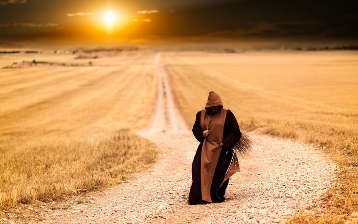 Monk walking on a dirt road through a wheat field at sunset