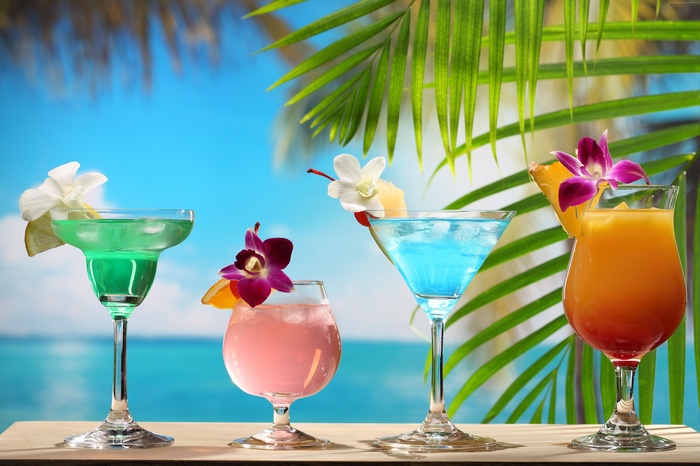 Four tropical cocktails on a wooden surface with palm trees and ocean background