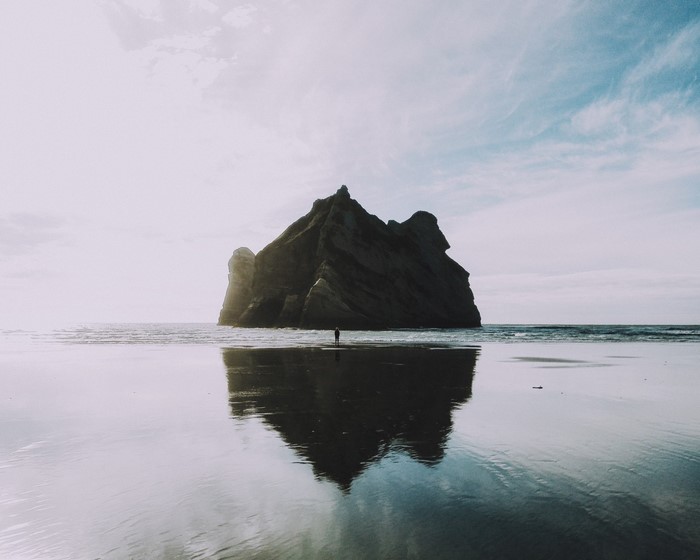 Lone figure on a beach with a large rock formation and reflection