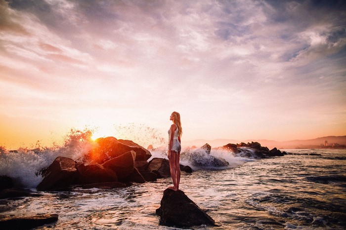 Woman standing on rock at sunset with waves crashing