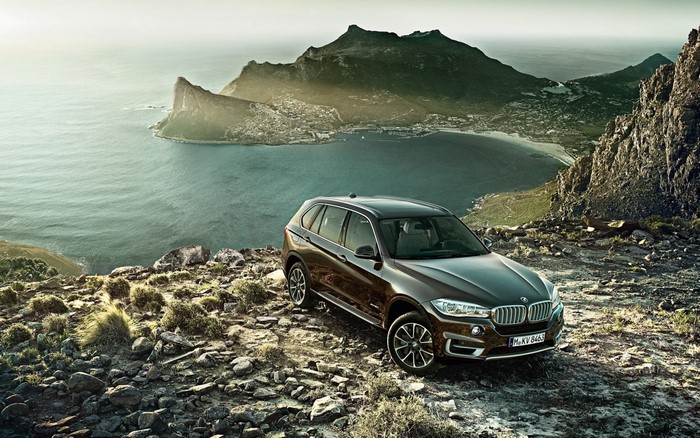 Brown BMW X5 SUV parked on rocky mountain overlooking ocean and town