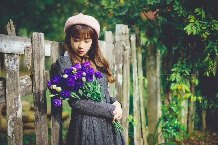 Young woman in a dress and beret holding flowers by a fence