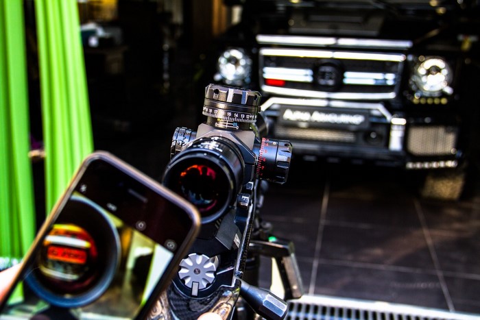 Sniper rifle scope and iPhone in front of a car