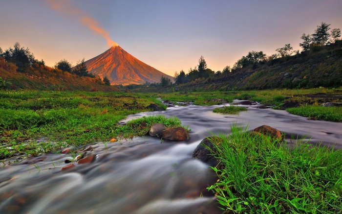 Volcano erupting with smoke over a stream and grassy landscape