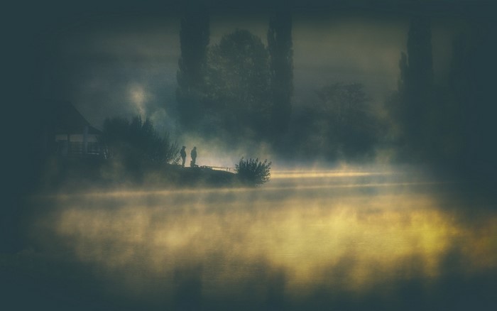 Two people stand by a misty lake at sunrise near a house