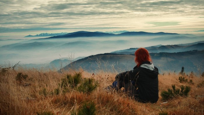 Woman with red hair sitting on a grassy hill overlooking misty mountains