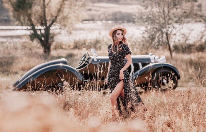 Woman in floral dress and hat posing by vintage car