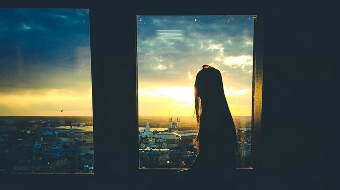 Silhouette of a woman looking at a city sunset through a window