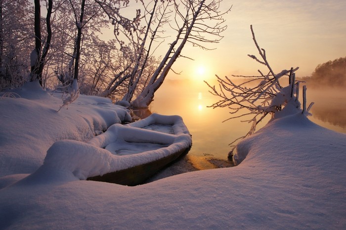 Snow-covered boat on a misty winter lake at sunrise