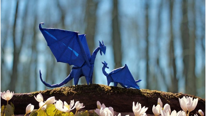 Two blue origami dragons on a mossy branch with white flowers