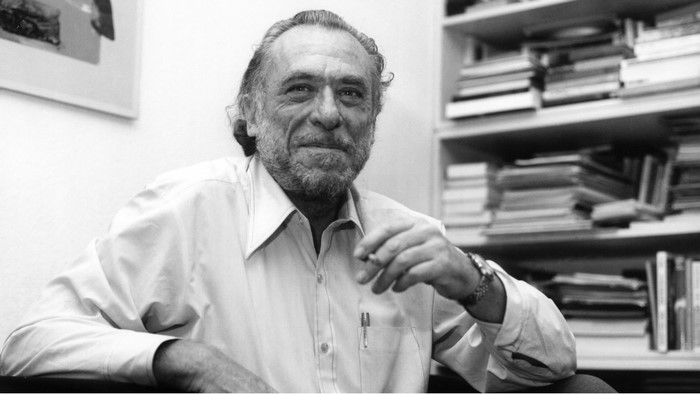 Charles Bukowski smoking a cigarette in front of bookshelves