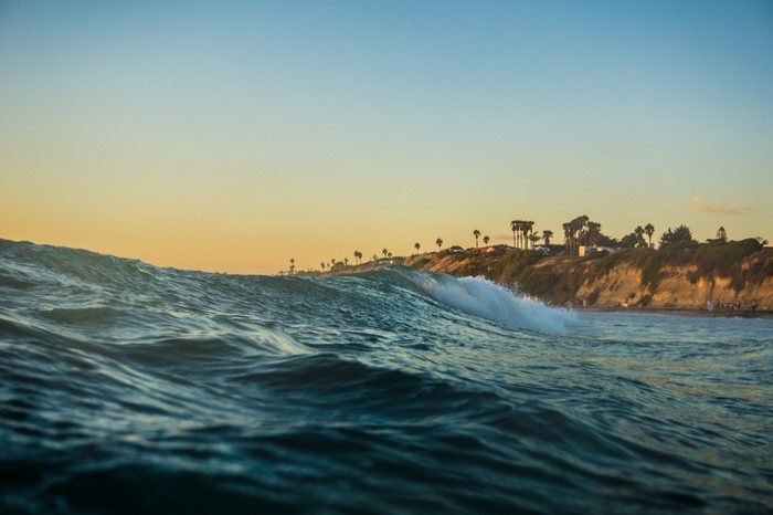 Ocean wave crashing near a cliffside with palm trees at sunset