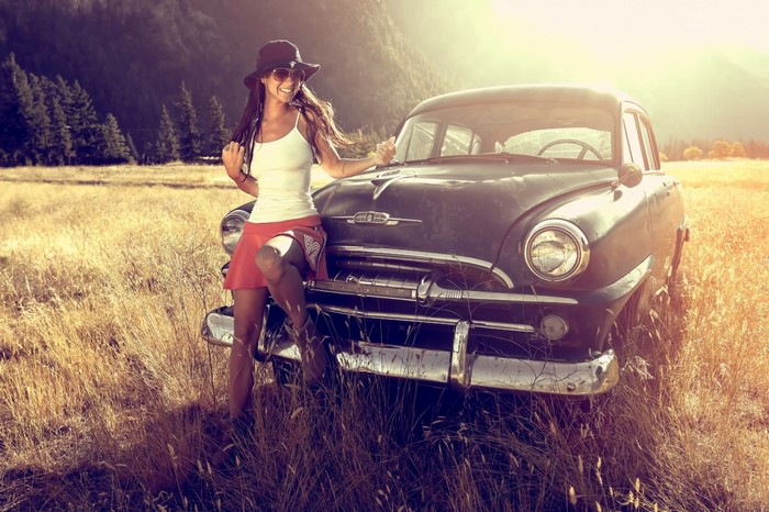 Woman in hat and skirt leaning on vintage car in field