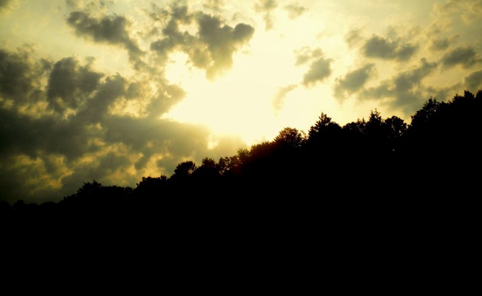 Sunlight filtering through clouds over silhouetted trees on a hill