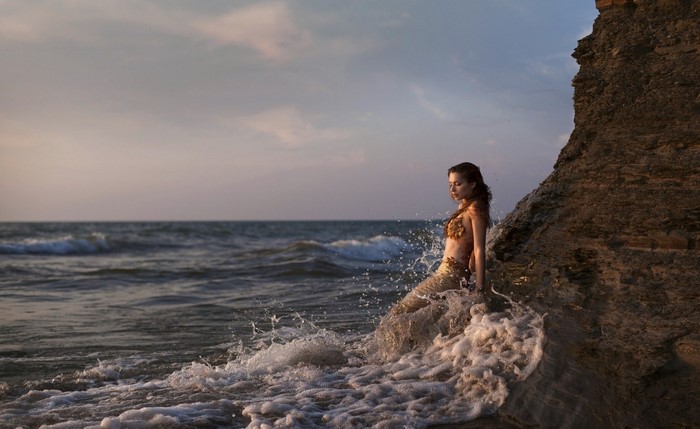 Woman in mermaid tail on rocky shore with waves