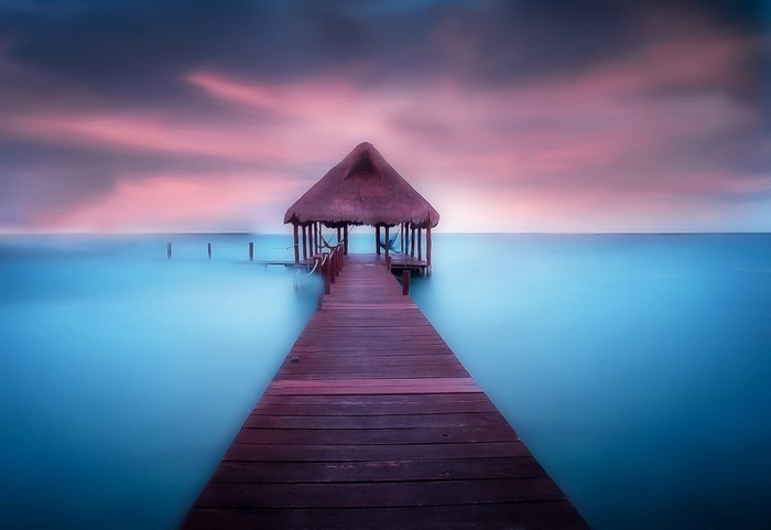 Wooden pier with thatched hut over calm ocean at sunset